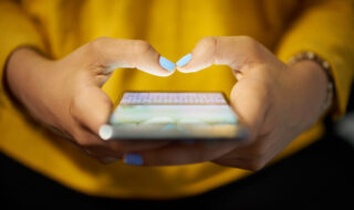 Young woman using cell phone to send text message on social network at night. Closeup of hands with computer laptop in background