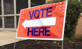 Large Orange background colored placard with Bold blue lettering that says Vote here