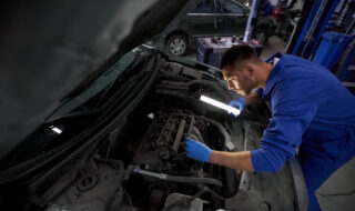a male mechanic with gloves looking under the hood of a blue care at night with a flashlight
