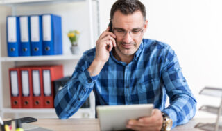 A man sitting down at a table on the cellphone with a tablet in his hand smiling.