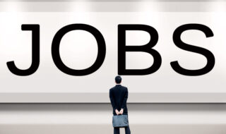 A man dressed in a black suit with a suitcase behind his back standing in front of a huge black and white sigh with the word JOBS in black font